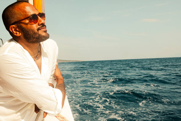 This image features a stylish, smiling man with a beard and sunglasses, wearing a white linen shirt and colourful beaded necklaces, sitting on a wooden boat during a sunny day at sea in Bodrum, Turkey. He is holding a smartphone and appears to be enjoying a relaxed moment, possibly taking a selfie or video calling. The background showcases the open ocean with gentle waves and a clear blue sky, with a distant coastline visible. The warm sunlight casts a golden glow on his face, enhancing the vacation, leisure, and travel vibes.
