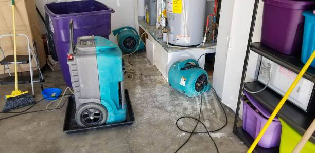 A garage undergoing water damage restoration with professional equipment, including dehumidifiers and air movers, actively drying the space near a water heater. The scene illustrates a thorough cleanup process to prevent mold and restore functionality. Ideal for showcasing emergency water damage services or professional restoration solutions.