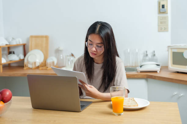 istock Asian woman in glasses reading documents while working on laptop in kitchen. Cozy home setting with orange juice, breakfast, and wooden decor creates a relaxed work-from-home environment. 2197167381