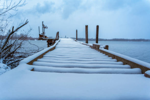 Snow-covered wooden dock extending into a calm winter lake under an overcast sky. Frosty and serene atmosphere in a natural, rustic setting. No people, peaceful scene