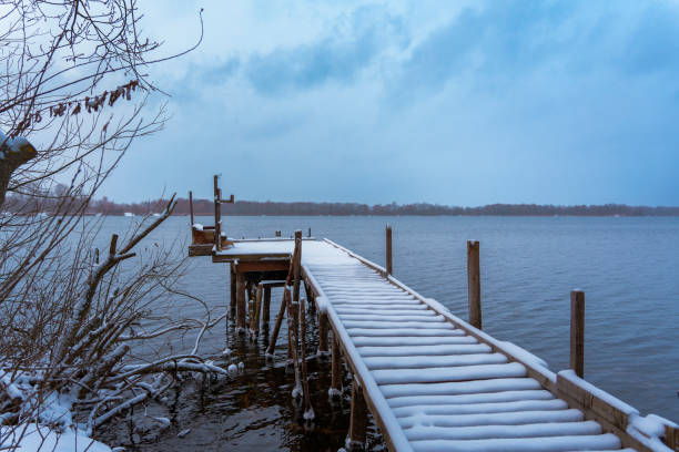 Snow-covered wooden pier extending over a tranquil winter lake. Overcast sky and frosty atmosphere create a serene and peaceful scene. Rustic and natural setting. No people