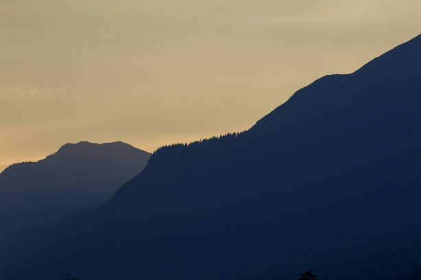 This image features two distinct mountain ranges silhouetted against a softly colored sky. The mountains are layered, with the closer range appearing darker and more prominent, partially obscuring the more distant range. The ridgelines are visible, creating a sense of depth and perspective. The sky is a gradient of light tones, suggesting either dawn or dusk, and provides a smooth backdrop for the dark, textured mountains. The overall composition is minimalist and serene, focusing on the shapes and layers of the mountains against the tranquil sky. A few small details are visible at the bottom of the frame, hinting at a landscape or vegetation in the foreground.