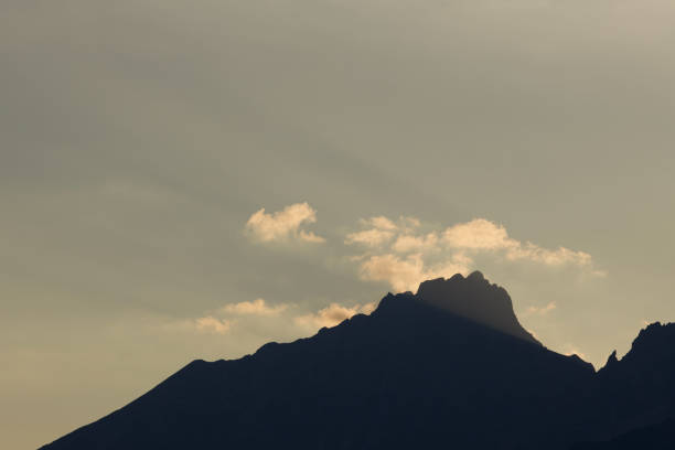 This image features the silhouette of a mountain peak with a unique, jagged profile against a muted, hazy sky. The mountain is dark, its details obscured by the lack of direct lighting, emphasizing its rugged shape. The sky behind the peak is a soft, uniform color, providing a subtle backdrop that allows the mountain's outline to stand out. Wispy, light clouds are scattered across the sky, partially obscuring the mountain and adding a touch of texture to the otherwise smooth background. The overall composition is minimalist, focusing on the contrast between the solid, dark mountain and the soft, light sky. The scene evokes a sense of quiet solitude and the remote beauty of a mountain landscape.