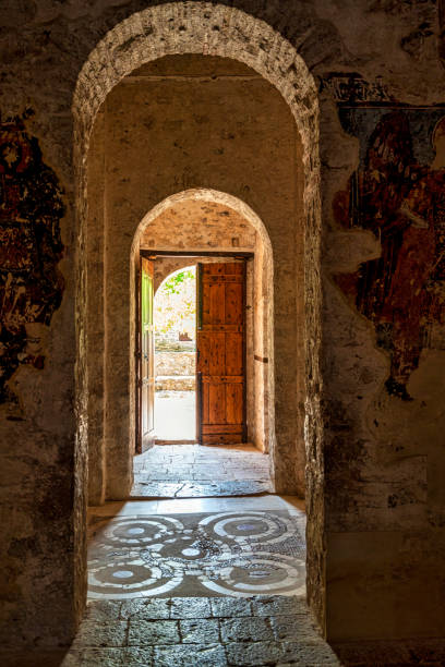 A historic stone archway inside Andromonastiro Monastery in Messinia, Greece, with an open wooden door leading to a sunlit courtyard.