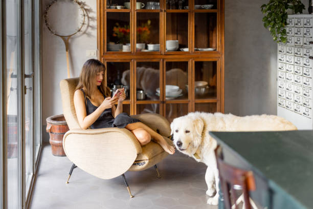 A woman sits in a stylish mid-century armchair, scrolling on her phone, while her large white dog stands nearby. The warm natural light and vintage wooden cabinet create a cozy and nostalgic atmosphere