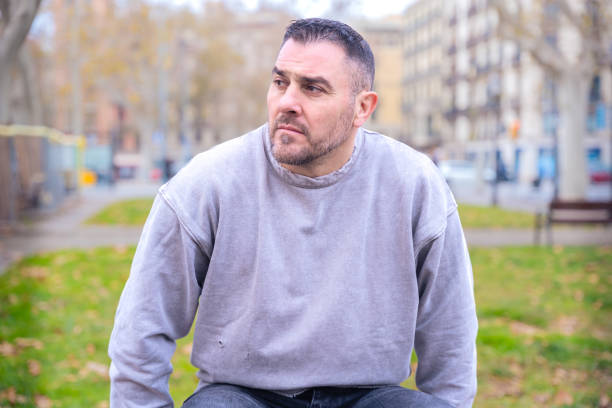 Man in a gray sweatshirt sitting on a bench in an urban park, looking thoughtful as he gazes into the distance. Background of trees, buildings, and an autumn city atmosphere.