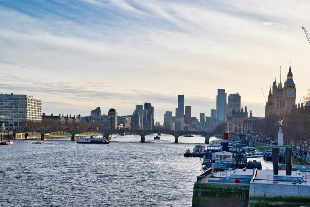 A wide shot of the River Thames in London, featuring Westminster Bridge, various boats, buildings including the Houses of Parliament and Big Ben, and a modern city skyline.  The sky is partly cloudy, with a soft light.