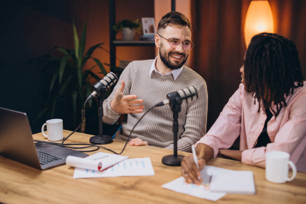 istock Two podcasters sitting at a wooden table, recording a podcast episode and taking notes, using professional microphones and a laptop 2197212331