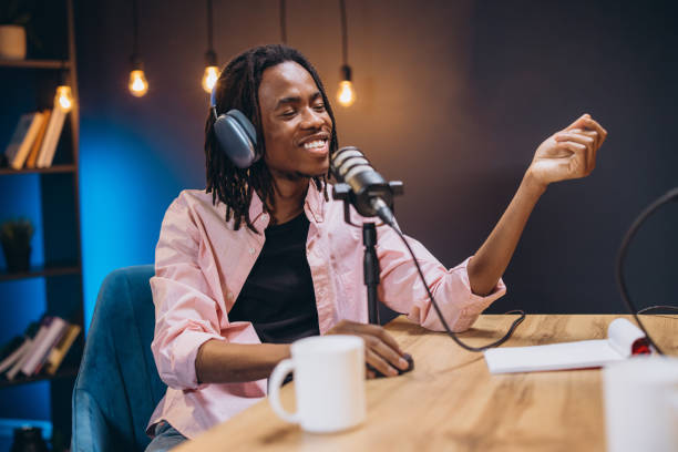 istock Radio host engaging with a microphone while recording a podcast in a modern home studio, surrounded by audio equipment and books 2197212761