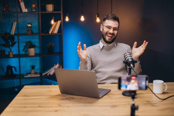 Happy male podcaster gesturing while recording audio podcast in his home studio using professional microphone and laptop