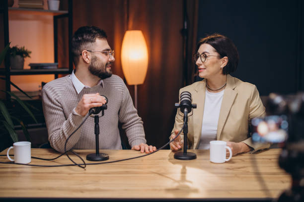 Podcasters recording radio show, speaking into professional microphones while camera captures collaborative media production moment