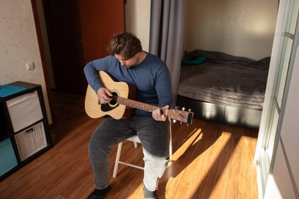istock Man Strumming Acoustic Guitar, sitting indoor practicing, learning musical instrument 2197233023