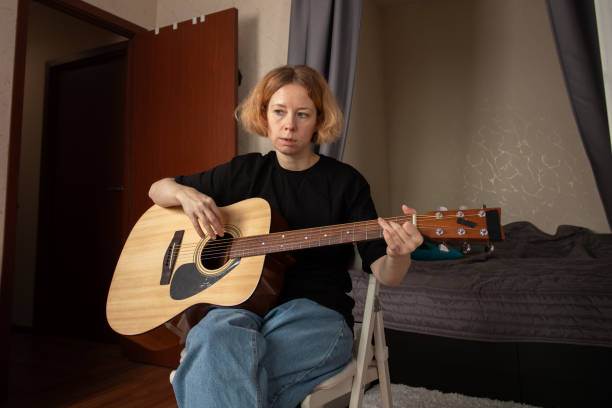 istock Woman Practicing Guitar in Cozy Bedroom, practicing chords on an acoustic guitar, focused on skill and education. 2197233112