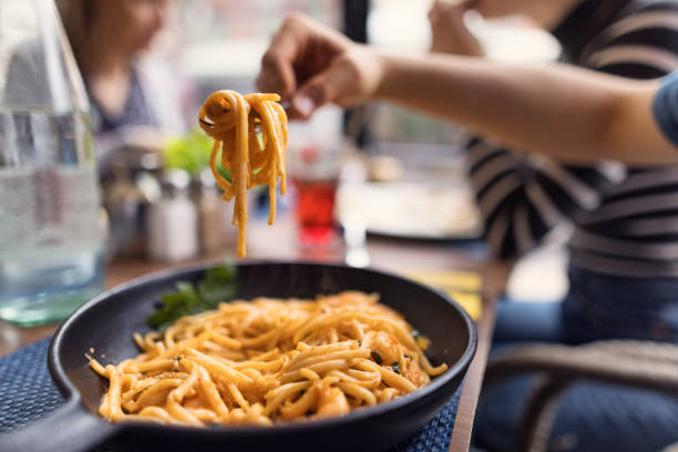 A close-up of a black skillet filled with steaming pasta, with a hand lifting a tasty forkful of noodles. The background shows a blurred restaurant setting with people sitting at the table, enjoying their meals. The table is set with glasses, utensils, and a water carafe.\nShot with Canon R5