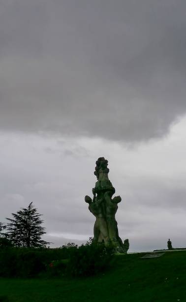 A dramatic shot of a monumental stone sculpture featuring intertwined human figures holding a symbolic structure at the top. The imposing artwork stands against a moody, overcast sky, enhancing its mysterious and powerful presence. The dark clouds add depth and contrast, making this image perfect for themes related to art, history, culture, and dramatic landscapes.