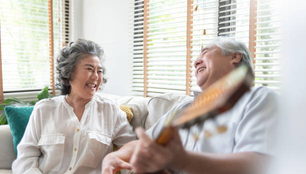 istock Asian elderly couple joyfully shares moment on sofa, with one playing guitar and both laughing, surrounded by natural light streaming through window blinds 2197243436