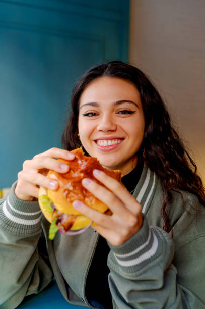 istock Happy young woman eating burger in restaurant 2197246950