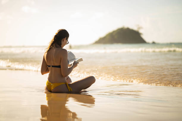 girl sits on the beach, captivated by a book as the sun sets behind distant hills, with gentle waves lapping at the shore creating a serene atmosphere.