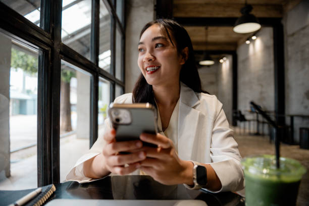istock Young asian businesswoman sitting in a cafe, using her smartphone while looking away with a cheerful smile. Enjoying the urban atmosphere and connecting with friends online 2197261731