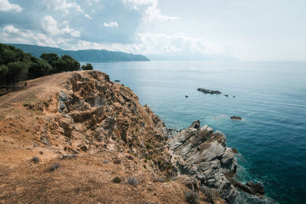 A scenic coastal landscape with rocky cliffs overlooking a calm sea. The sky is partly cloudy, and the distant horizon is visible. The vegetation is sparse, with some trees on the cliff.