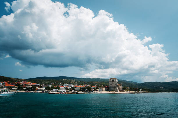 A scenic view of a coastal town with a tower, surrounded by mountains and a clear blue sky with fluffy clouds. The water is calm and reflects the landscape.