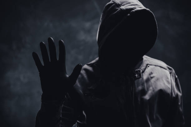 teenage boy wearing a black hood over his head so his face is not visible, standing sideways against a mesh fence, eyes looking down. Concept of backlit image, teenagers, hackers.