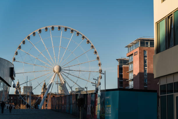 Liverpool, UK, January 25th 2025. Ferris wheel beside modern buildings and pathway under a clear blue sky.