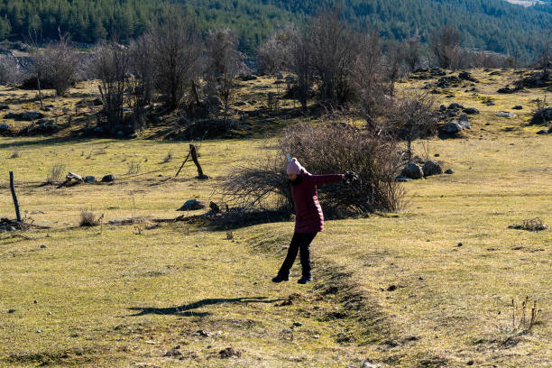 Having moved away from the city to experience the tranquility of nature, the woman walks past two bare trees, with a high, densely forested hill rising behind her.