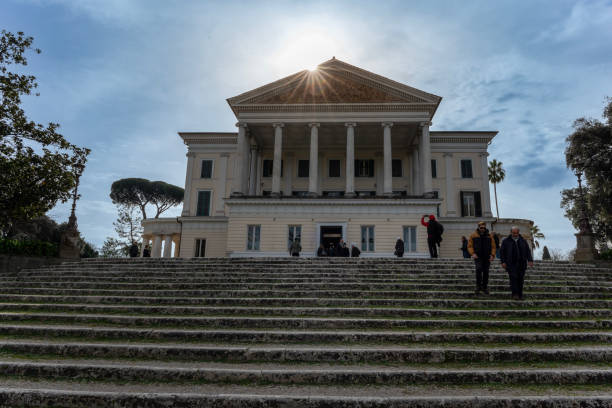 A warm-toned building surrounded by lush greenery, tall trees, and a serene blue sky in Rome, Italy.
