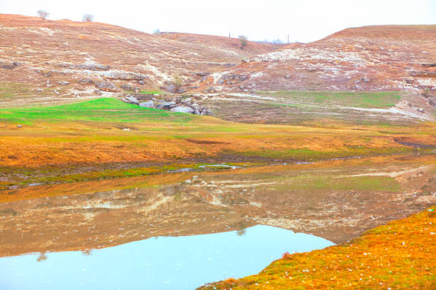 Serene landscape with a small body of water reflecting the surrounding hills and sparse vegetation. Scene is characterized by a mix of green patches of grass, with rocky outcrops visible on the hills