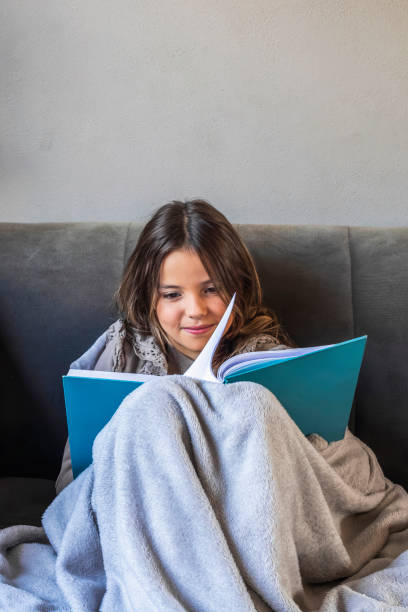 istock Girl reading a book wrapped in a blanket on sofa at home 2197303992