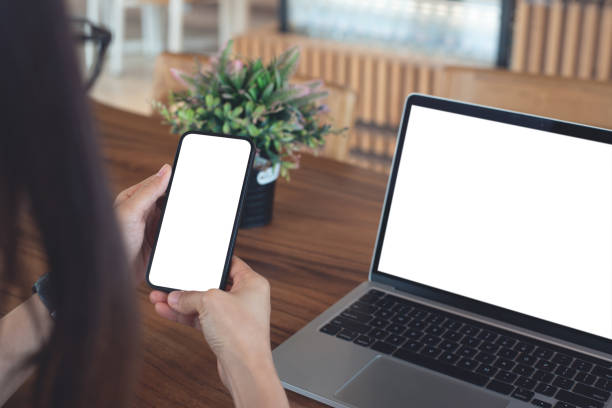 istock Mockup blank white screen cell phone for web template or mobile app design. Woman hand using mobile phone during working on laptop computer on wooden table at coffee shop or home office 2197327971