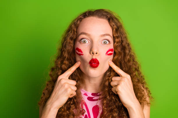 istock Young woman with red lipstick kissing marks on her cheeks against a solid green background 2197359703