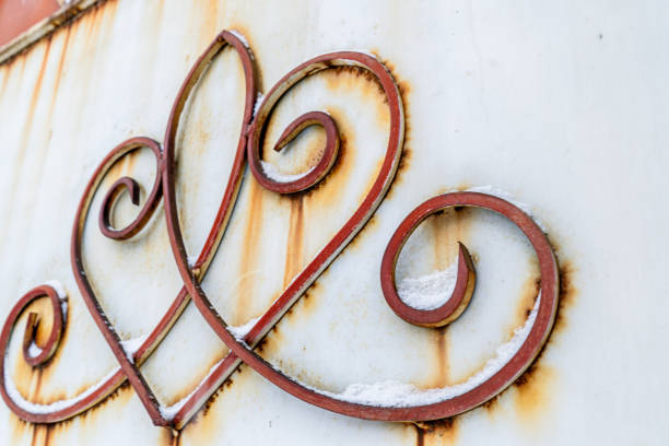 An intricate wrought iron design with elegant swirling patterns is attached to a white metal surface, now weathered by time. Rust stains bleed through, contrasting with the once-clean background, while patches of snow settle delicately on the curves of the metalwork.