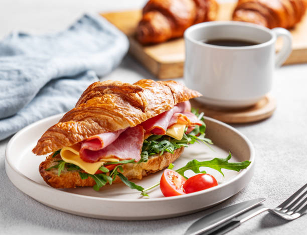 Croissant sandwich with ham, cheese, tomato  and arugula on a plate on a white background