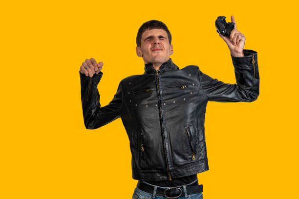 Young man wearing black leather jacket holding broken controller celebrating victory on yellow background