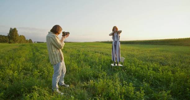 A man takes pictures of a young woman at sunset in a field. A woman poses standing on a chair