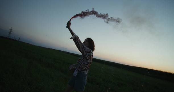 A young woman blows pink smoke standing in a field on a summer evening. She smiles and spins around