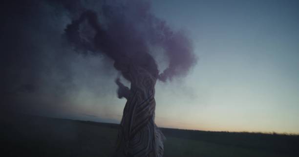 A young woman blows pink and blue smoke running across a field on a summer evening