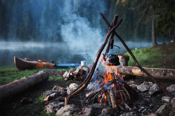 An old-fashioned teapot steaming over an open flame at a campsite, with a picturesque lake view in the distance.
