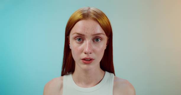 A woman puts a cosmetic mask on her face. She smiles at the camera while standing in the studio background