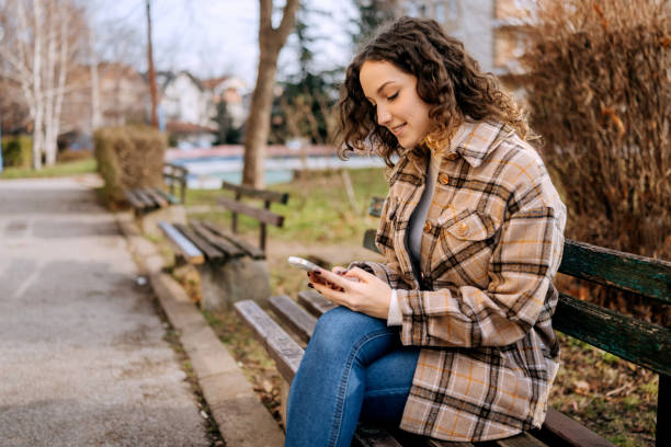 istock Young beautiful woman using smart phone while sitting on park bench outdoors 2197392273