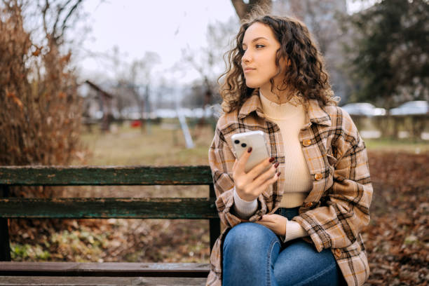 istock Young beautiful woman using smart phone while sitting on park bench outdoors 2197392284