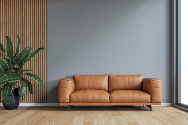This image features a modern, minimalist living space with a brown leather sofa placed against a muted gray wall. To the left, a wooden slat accent wall adds texture, accompanied by a lush green plant in a black pot (a strelitzia / bird of paradise plant). Natural light from a large window on the right enhances the warm tones of the wooden flooring, creating a cozy and elegant atmosphere. 3D rendered image.