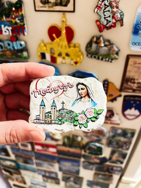 A hand holds a colorful souvenir magnet displaying Medjugorje scenery and a Virgin Mary depiction, surrounded by a collage of other travel magnets.