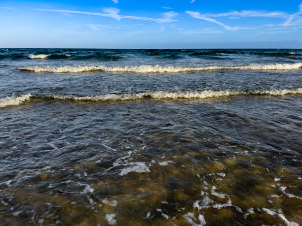 A serene view of shallow tidal pools at Lauderdale-by-the-Sea Beach, Florida, capturing the intricate patterns of light and shadow on the sandy ocean floor. The crystal-clear water ripples gently, reflecting the blue sky with scattered wispy clouds. Ocean waves roll in the background, creating a peaceful and tropical coastal atmosphere.