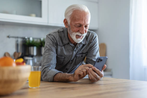 istock Mature man standing alone in kitchen, drinking orange juice and using smart phone. Elderly man, pensioner, sitting in kitchen and using mobile phone, he is in nursing home or in his apartment, concept of aging and happy retirement days. 2197410156