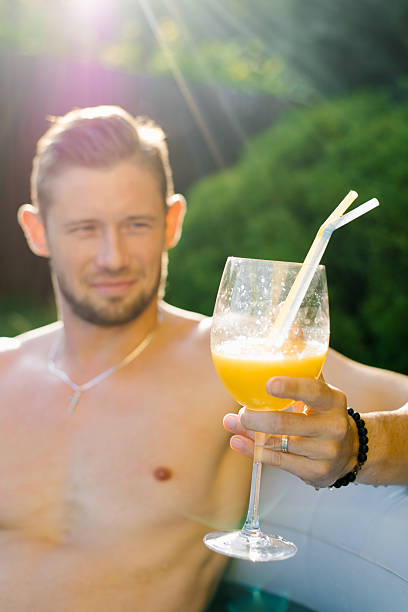 A relaxed man sitting in a pool, holding a glass of orange juice with a straw, surrounded by lush greenery and enjoying a peaceful moment.