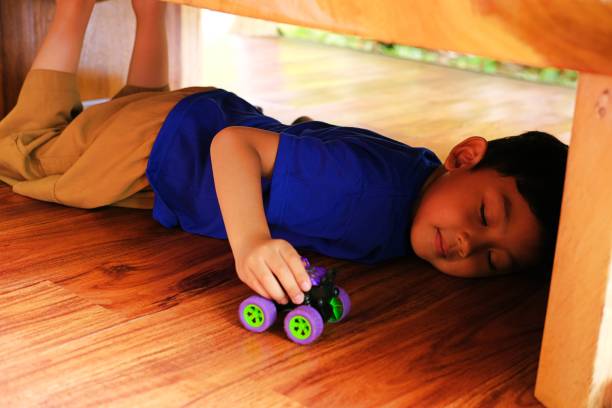 istock A young boy plays joyfully under a wooden table with a small toy car. 2197418595