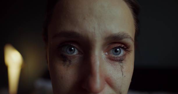A woman is crying sitting on a bed at home. Her mascara was leaking. She's looking at the camera. Close-up of her face